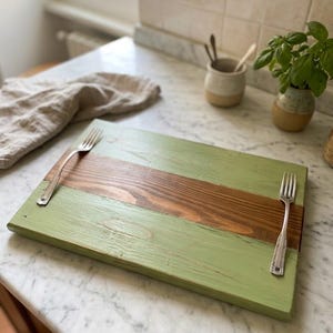 Könnte beinhalten: Ein rechteckiges Servierbrett aus Holz mit einem horizontalen Mittelstreifen aus naturbelassenem, dunkelbraunem Holz, flankiert von zwei Abschnitten aus hell salbeigrün lackiertem Holz, mit zwei silberfarbenen Metallgabeln darauf.