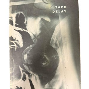 Può includere: Una copertina di libro in bianco e nero con la scritta bianca "TAPE DELAY". L'immagine presenta una figura sfocata e un registratore a bobine. Il design ha un'atmosfera vintage, che ricorda la vecchia tecnologia audio.