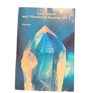 May include: A book titled "Gem Elixirs and Vibrational Healing, Vol. I" by Gurudas. The cover features a blue and turquoise crystal illustration against a dark blue background. The book has a worn appearance.
