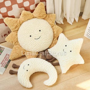 May include: A set of three plush cushions featuring a sun with golden yellow rays, a white star, and a white crescent moon. Each cushion has a textured, wavy fabric surface, a simple embroidered smiling face, and small brown dangling legs.