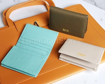 May include: Three leather card holders with personalized initials. The card holders are in shades of green, blue, and beige. The card holders are on top of a brown leather tote bag.