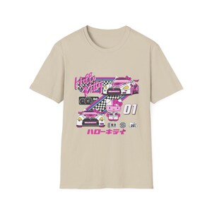 Op de afbeelding: Een beige T-shirt met een Hello Kitty race-thema. Het ontwerp toont Hello Kitty in een roze racepak, rijdend in een witte raceauto met roze accenten. De tekst "Hello Kitty" is in roze en wit, en de tekst "ハローキティ" is in roze.