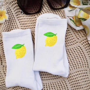 Könnte beinhalten: Ein Paar weiße Socken mit gestickten gelben Zitronen und grünen Blättern. Eine schwarze Sonnenbrille mit Schachbrettmuster an den Bügeln und ein Schal mit Zitronenmuster sind ebenfalls zu sehen. Die Artikel sind auf einer beigen Strickoberfläche angeordnet.