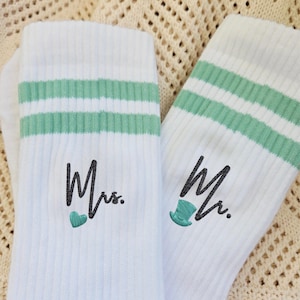 Könnte beinhalten: Paar weiße Socken mit mintgrünen Streifen am oberen Rand. Eine Socke ist mit dem Wort "Mrs." mit einem Herz bestickt, die andere mit "Mr." mit einem Zylinder. Die Socken liegen auf einer cremefarbenen, strukturierten Oberfläche.