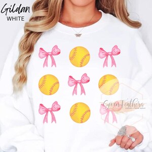 May include: White Gildan sweatshirt with a softball and pink bow design. The design features six softballs and six bows.
