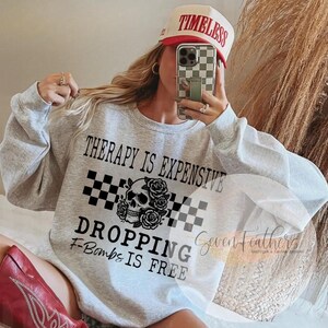 May include: A light gray sweatshirt with the text "THERAPY IS EXPENSIVE DROPPING F-BOMBS IS FREE" and a skull graphic with roses. The image also shows a white and red baseball cap that says "TIMELESS".