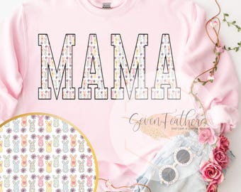 Puede incluir: Sudadera rosa con la palabra "MAMA" en letras negras delineadas. Las letras están rellenas con un patrón de conejos y flores de color pastel. La sudadera está sobre una superficie blanca con un par de gafas de sol blancas y flores rosas.