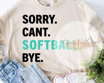 May include: A beige sweatshirt with the text "Sorry. Cant. Softball Bye." in black and teal lettering. The sweatshirt is laid out on a surface with blue jeans and white sneakers.
