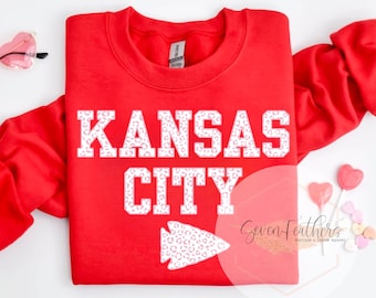 May include: Red sweatshirt with white text that says "Kansas City" with a pink and white leopard print design. The text and arrow are outlined in white.