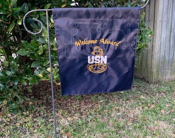 Puede incluir: Una bandera de jardín negra con el texto "Welcome Aboard" y un ancla bordada en oro con las letras "USN" en blanco. La bandera está colgada de un poste de metal negro.