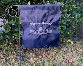 Puede incluir: Una bandera de jardín azul marino con las palabras "Welcome Aboard" en escritura blanca sobre un emblema bordado blanco. La bandera cuelga de una estaca de jardín de metal frente a un arbusto verde.