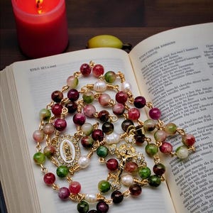 May include: A rosary with colorful gemstone beads, interspersed with pearls, and gold-toned chain. The rosary features a gold crucifix and a pendant with a religious image. A lit red candle and an open book are in the background.