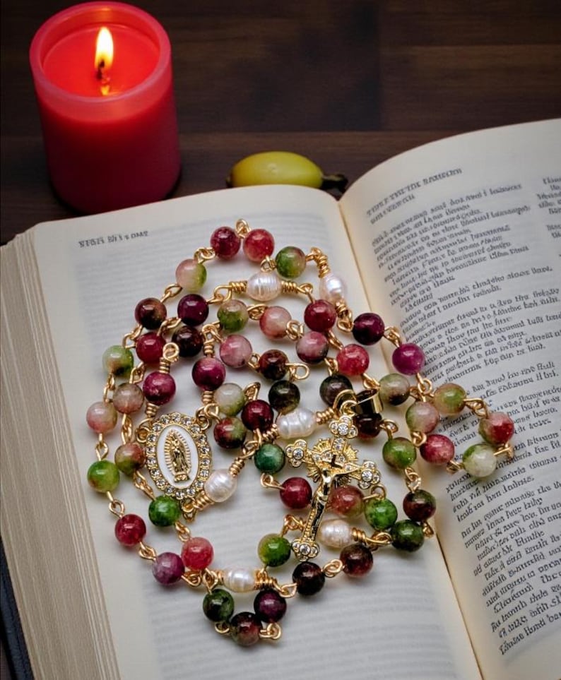 May include: A rosary with colorful gemstone beads, interspersed with pearls, and gold-toned chain. The rosary features a gold crucifix and a pendant with a religious image. A lit red candle and an open book are in the background.