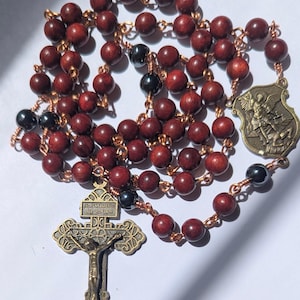 May include: A rosary with dark red wooden beads and black beads, strung on copper wire. The rosary features a bronze crucifix and a shield-shaped medal with an image of a saint. The crucifix has the letters INRI and other text.