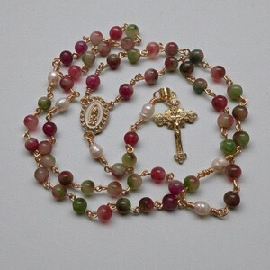 May include: A rosary necklace with multi-colored beads in shades of red, green, and brown, interspersed with white pearls. The gold-toned chain features a gold cross pendant and a decorative centerpiece with an oval design.