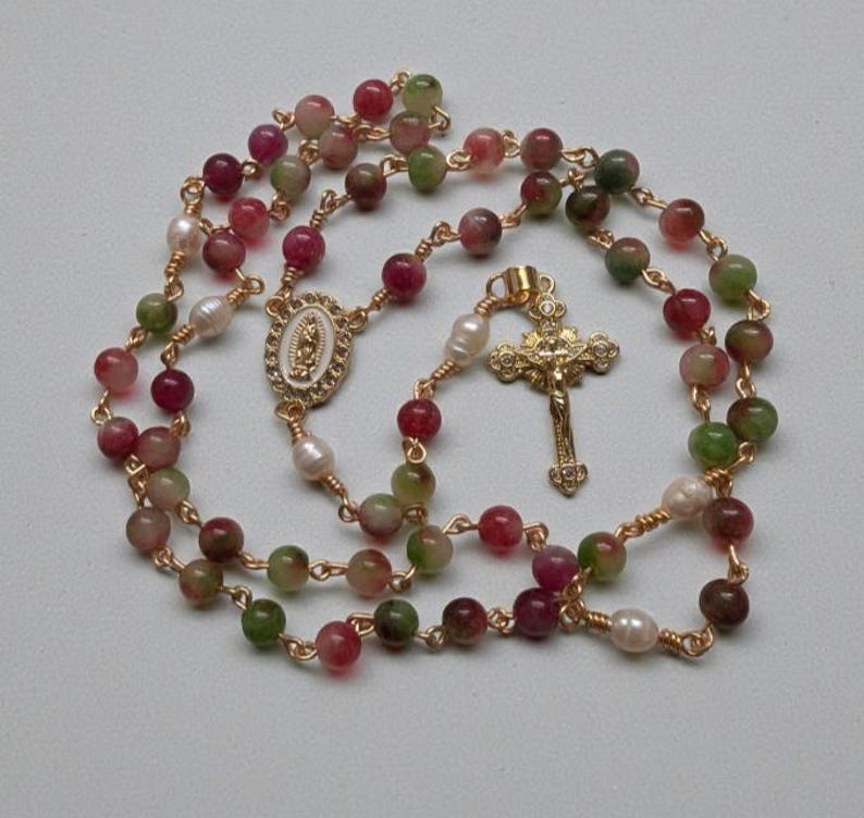 May include: A rosary necklace with multi-colored beads in shades of red, green, and brown, interspersed with white pearls. The gold-toned chain features a gold cross pendant and a decorative centerpiece with an oval design.