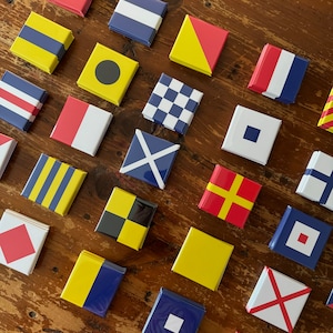May include: A collection of colorful nautical signal flags with various letters and symbols. The flags are made of fabric and have a white background with different colored designs. The flags are arranged in a grid pattern on a wooden surface.