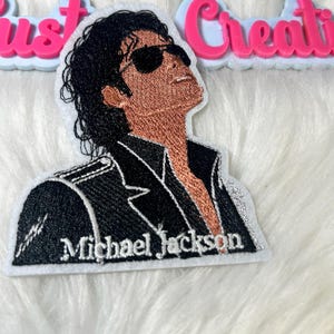 May include: Embroidered patch featuring a portrait of Michael Jackson wearing sunglasses, looking upwards. The patch has a black jacket with white stitching and the name "Michael Jackson" embroidered in white.