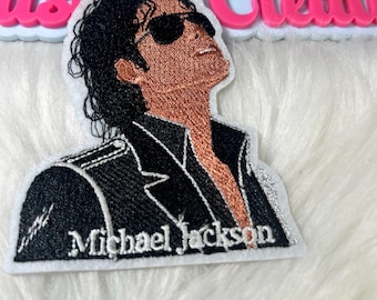 May include: Embroidered patch featuring a portrait of Michael Jackson wearing sunglasses, looking upwards. The patch has a black jacket with white stitching and the name "Michael Jackson" embroidered in white.