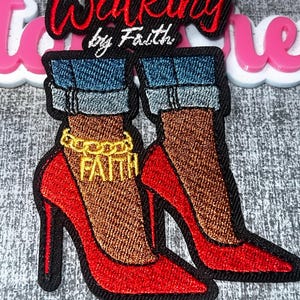 Pode incluir: Um emblema bordado com o texto Walking by Faith em fio vermelho e branco. O design mostra duas pernas vestindo calças jeans azuis com bainha dobrada e sapatos de salto alto vermelhos. Um tornozelo tem uma corrente dourada com a palavra FAITH.