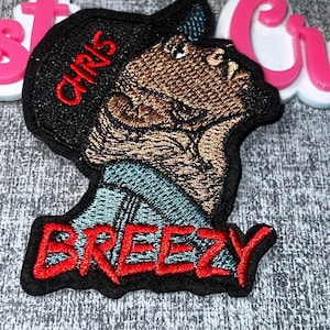 May include: Embroidered patch featuring a brown and black cartoon character with the name "CHRIS" in red on a black hat. The word "BREEZY" is also in red on the bottom of the patch. The patch has a black border and a blue and black collar.