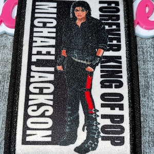 以下が含まれることがあります： マイケル・ジャクソンの肖像画が描かれた長方形のパッチ。パッチには、左側に縦書きで「MICHAEL JACKSON」、右側に「FOREVER KING OF POP」と書かれています。ジャクソンは黒い衣装に身を包み、ズボンには赤いストライプが入っています。