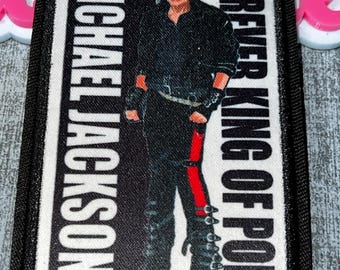 May include: A rectangular patch featuring a portrait of Michael Jackson. The patch has the text "MICHAEL JACKSON" vertically on the left and "FOREVER KING OF POP" on the right. Jackson is depicted in a black outfit with a red stripe on his pants.