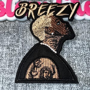 May include: Embroidered patch featuring a profile portrait with a black background and a tan and brown face. The word "Breezy" is embroidered in gold thread above the portrait. The patch is likely for clothing or accessories.