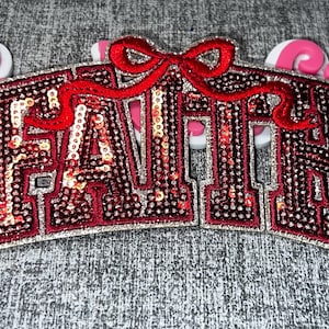 May include: A decorative patch featuring the word FAITH in bold, block-style letters. The letters are filled with dark red sequins and outlined in silver, topped with a prominent red sequined bow. The design is mounted on a white backing.