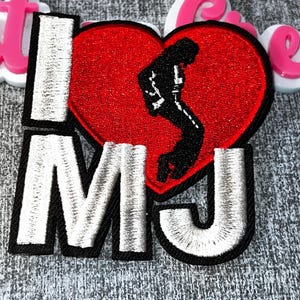 Può includere: Toppa ricamata con la scritta "I MJ" in bianco, con un cuore rosso raffigurante una silhouette nera di un ballerino. La toppa ha un bordo nero ed è probabilmente per abbigliamento o accessori.
