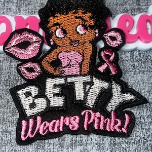 Puede incluir: Un parche bordado que presenta un personaje de dibujos animados con cabello negro y vestido rosa, acompañado de dos iconos de labios rosas y un lazo rosa. El texto dice BETTY en letras blancas y Wears Pink! en letra cursiva rosa.