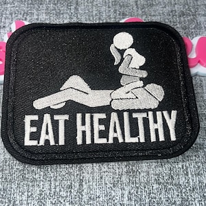 May include: A rectangular black embroidered patch featuring a white graphic of two figures in a sexual position with the text EAT HEALTHY in white capital letters at the bottom. The patch has a thick black border and rounded corners.