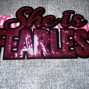 May include: A decorative patch featuring the text She is FEARLESS in dark magenta sequins with a black border. The word FEARLESS is in large block letters, while She is is in a cursive script. A partial pink and white text element is visible.
