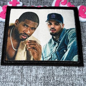 May include: A rectangular patch with two men depicted. One is wearing a beige top and a pearl necklace, holding a small object. The other is in a denim jacket and a dark blue cap. The patch has a black border.