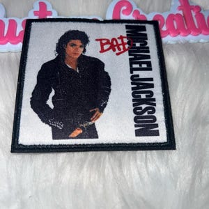 May include: A square patch featuring a portrait of Michael Jackson in a black jacket. The word "BAD" is printed in red diagonally across the image. The text "MICHAEL JACKSON" is printed vertically on the right side of the patch.