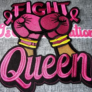 May include: An embroidered patch featuring two pink boxing gloves with yellow wrist wraps, positioned above the word Queen in pink script. The word Fight is embroidered in pink above the gloves, flanked by two pink awareness ribbons on a black background.