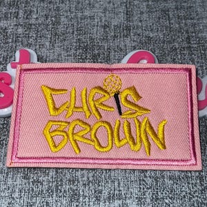 Puede incluir: Un parche rectangular rosa con las palabras "CHRIS BROWN" bordadas en hilo dorado. El parche tiene un diseño de micrófono dorado y un borde rosa.