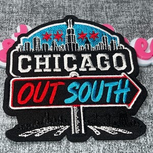 May include: A black embroidered patch featuring a Chicago skyline silhouette with red stars against a blue background. Below, a sign reads CHICAGO in white, and a directional arrow sign displays the red text OUT and blue text SOUTH.