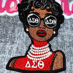 May include: Embroidered patch featuring a woman with dark skin, wearing round glasses with "ΔΣΘ" in white. She has black curly hair, red lips, and a red top with "ΔΣΘ". A white beaded necklace is also visible. The patch is outlined in black.