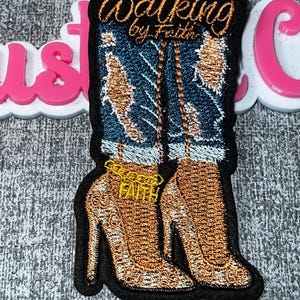 Puede incluir: Un parche bordado que muestra un par de piernas con jeans azules desgastados y zapatos de tacón dorados. Se ve una tobillera de cadena dorada con la palabra FAITH. El texto Walking by Faith está bordado en hilo de color cobre arriba.