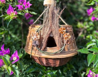 May include: A handcrafted birdhouse made of woven wicker and natural materials, suspended by a twine cord. The birdhouse features a conical roof of dried grasses and is adorned with decorative dried flowers. The background is filled with green foliage and vibrant purple flowers.