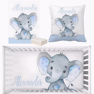 Puede incluir: Un juego de ropa de cama para bebé con una ilustración de acuarela de un elefante gris con detalles en azul. El juego incluye una sábana, una almohada, una manta y una manta doblada. El nombre "Alexander" está escrito en cursiva.