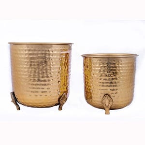 May include: Two gold-colored metal planters with a hammered texture. The larger planter has three decorative feet. The planters are cylindrical with a rolled rim. The planters are suitable for indoor or outdoor use.
