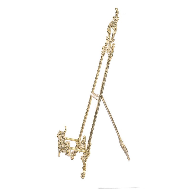 May include: A gold-colored decorative easel with ornate scrollwork. The easel has a three-legged design, with a support bar and a decorative top. The easel is designed to hold a picture frame or artwork.