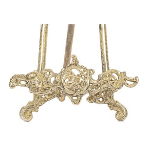 May include: A gold-colored, ornate easel with a decorative base and detailed scrollwork. The easel has a three-legged design and is made of metal. The intricate details and vintage style make it suitable for displaying artwork or photographs.