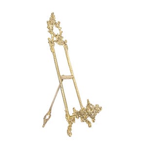 May include: A gold-colored, ornate easel with a decorative design. The easel has an A-frame structure with intricate scrollwork and flourishes. It is designed to hold a picture or artwork.