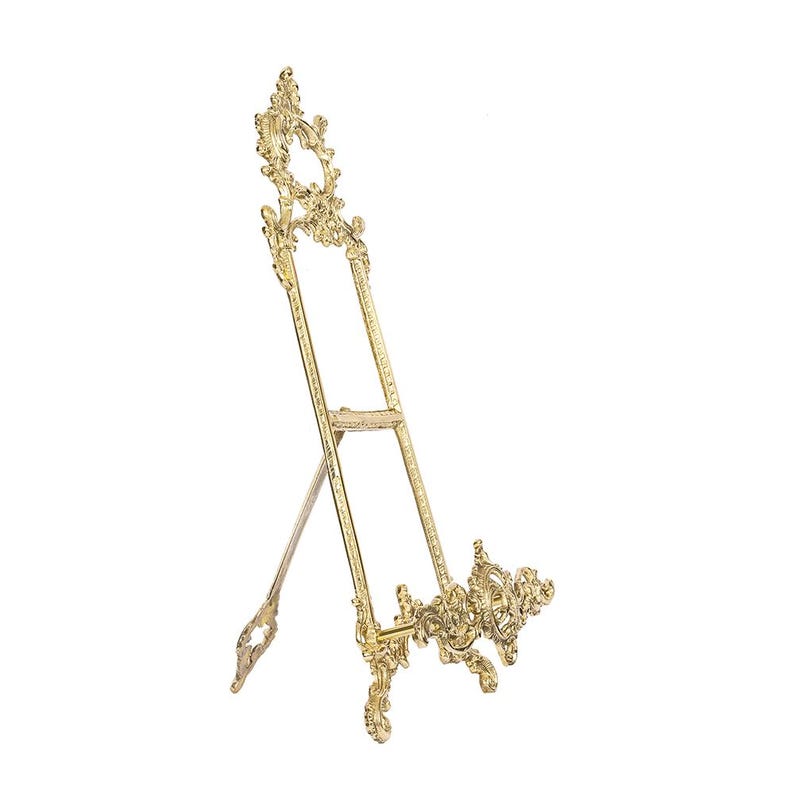 May include: A gold-colored, ornate easel with a decorative design. The easel has an A-frame structure with intricate scrollwork and flourishes. It is designed to hold a picture or artwork.