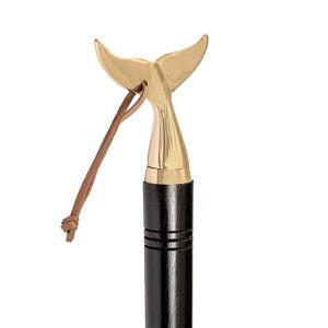 Pode incluir: Um bastão decorativo com uma pega de cauda de baleia em latão. A pega é fixada a um eixo de madeira escura com uma correia de couro. A cauda de baleia é dourada e o eixo é preto.