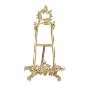 May include: A gold-colored, ornate easel with a decorative floral design. The easel has three legs and a frame to hold a picture or artwork. The top of the easel features a circular design with floral accents.