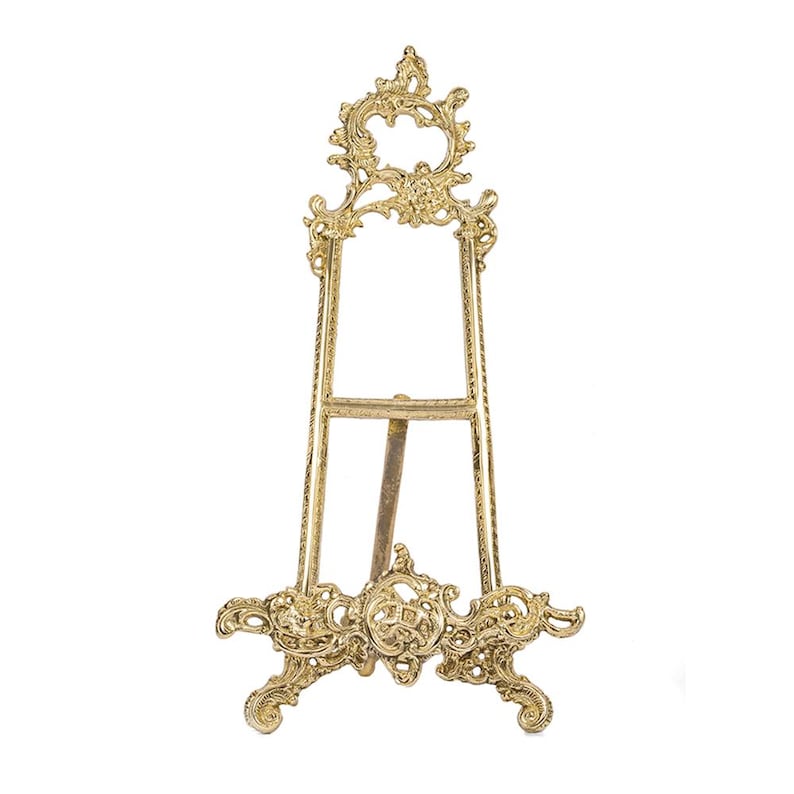 May include: A gold-colored, ornate easel with a decorative floral design. The easel has three legs and a frame to hold a picture or artwork. The top of the easel features a circular design with floral accents.